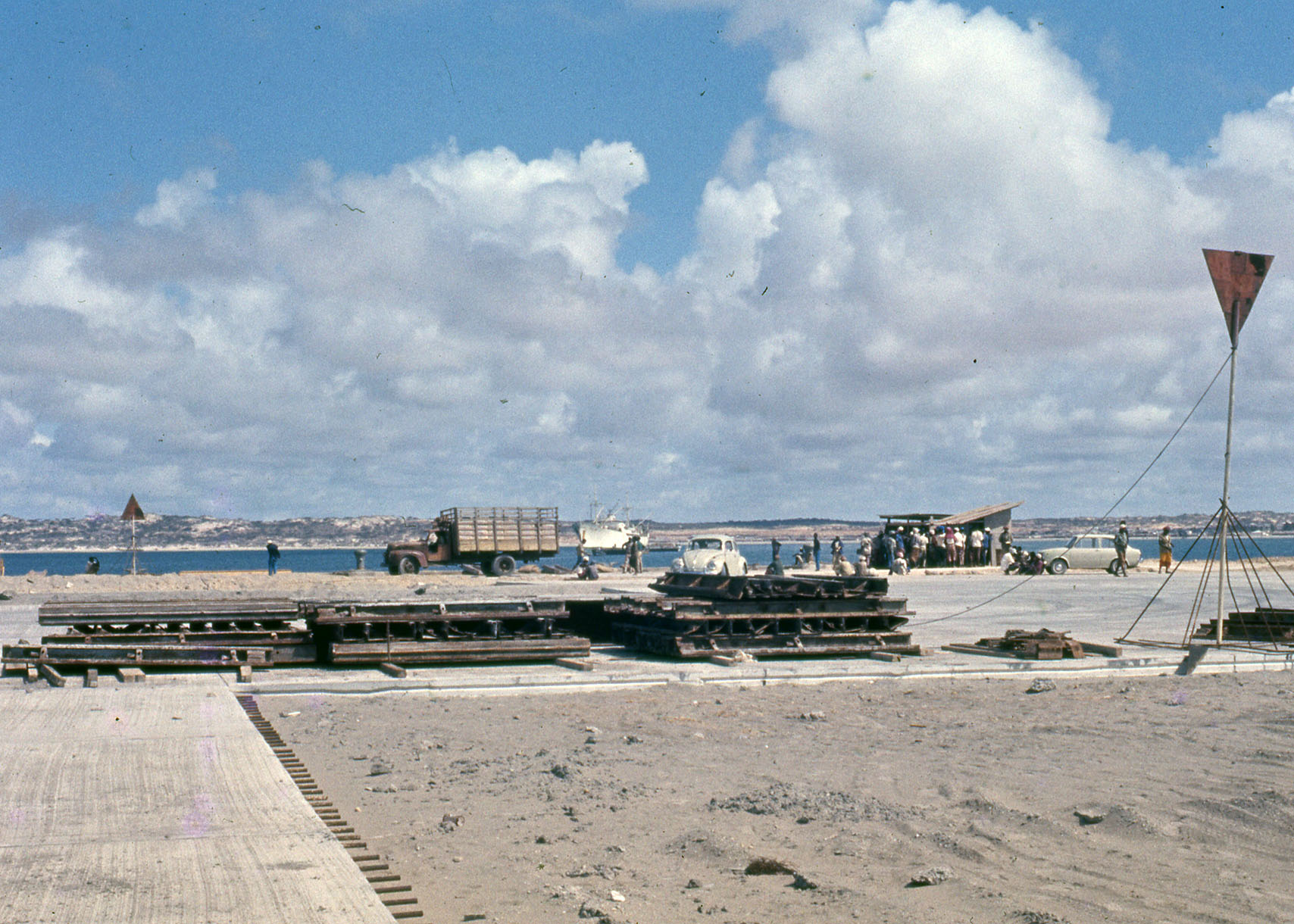 cars, trucks, construction materials, and people on a flat concrete pad near the waterside cars, trucks, construction materials, and people on a flat concrete pad near the waterside