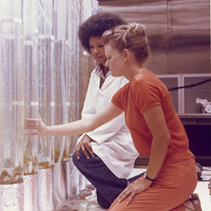 Two women kneeling in a plant laboratory with large water-filled tubes