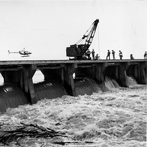 Men and crane atop large flood control structure with open spillways